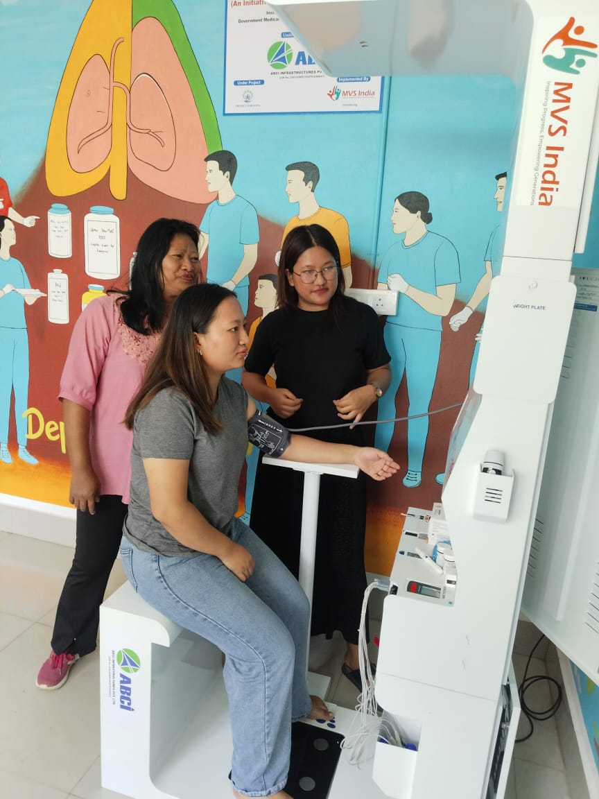 Woman seated at health kiosk with blood pressure cuff on arm, two women observing beside mural wall