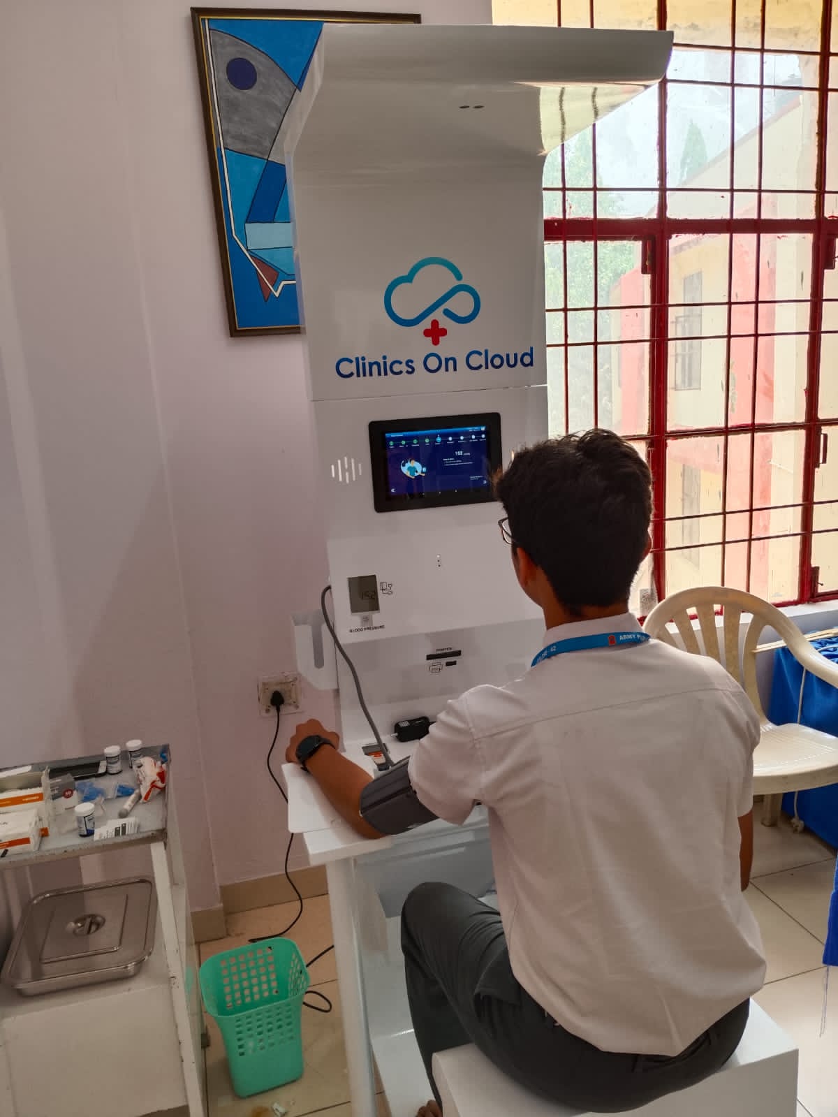 Student in uniform seated at a Clinics On Cloud health kiosk with arm in blood pressure cuff, near a clinic room window.