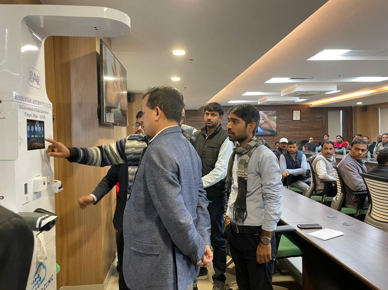 Man touches health kiosk screen from Hindustan Antibiotics Ltd as others watch in a boardroom with seated attendees.