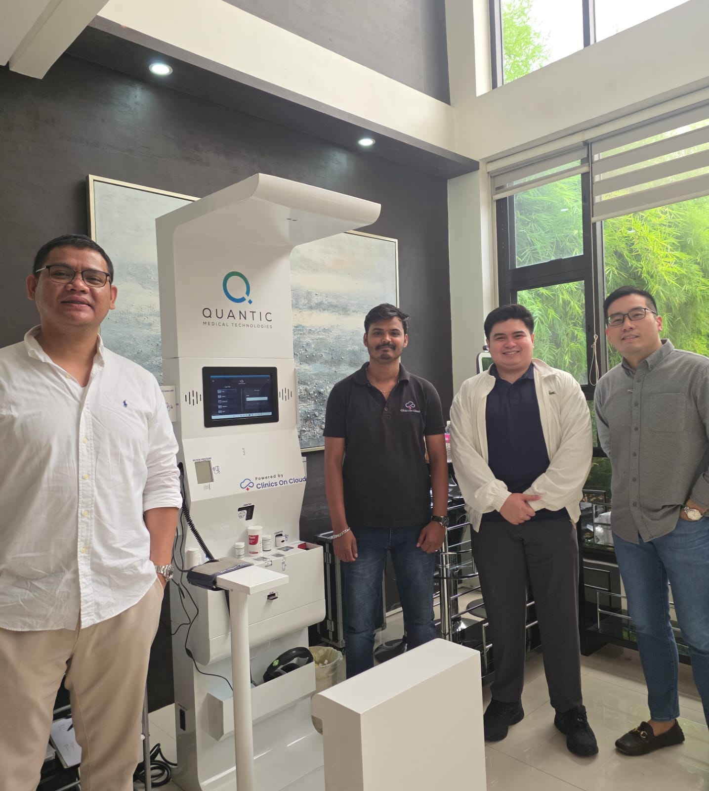 Four men stand beside a Quantic health kiosk with touchscreen in a bright indoor space with tall windows.