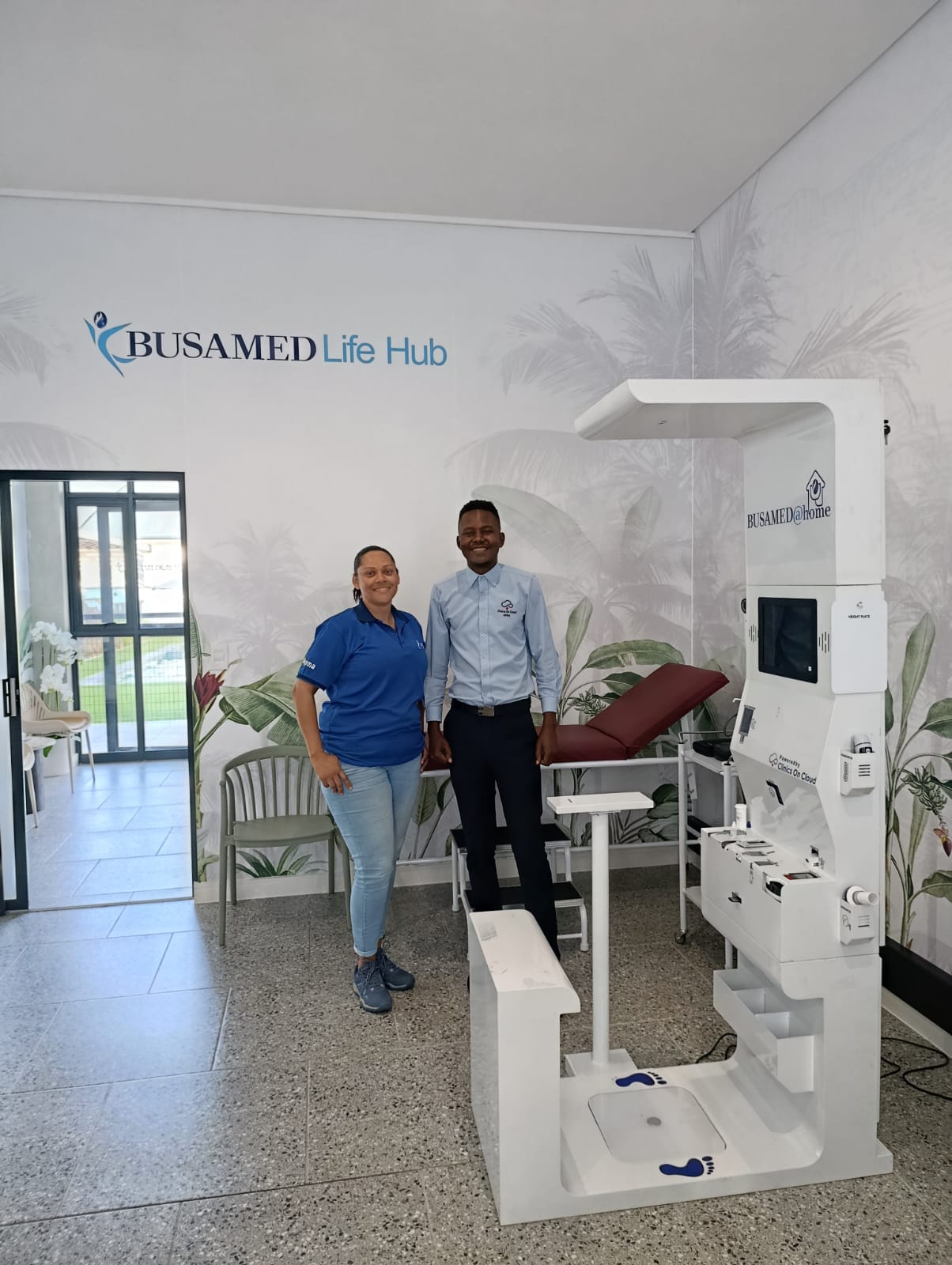 Two staff stand beside a Busamed@home health kiosk in the Busamed Life Hub room with palm-leaf wallpaper.