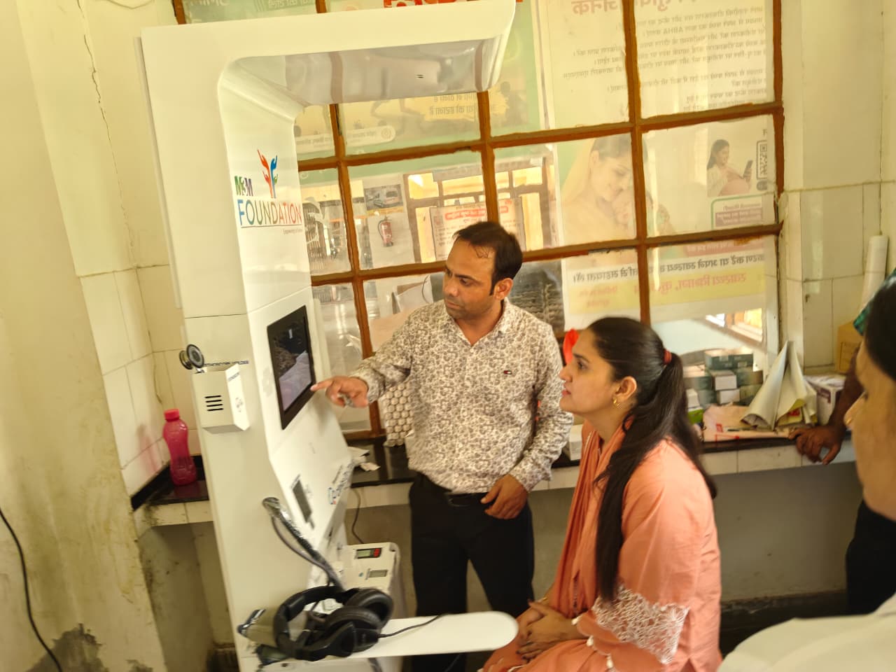 Man points at a health kiosk screen while a seated woman looks on inside a clinic room with health posters on the wall.
