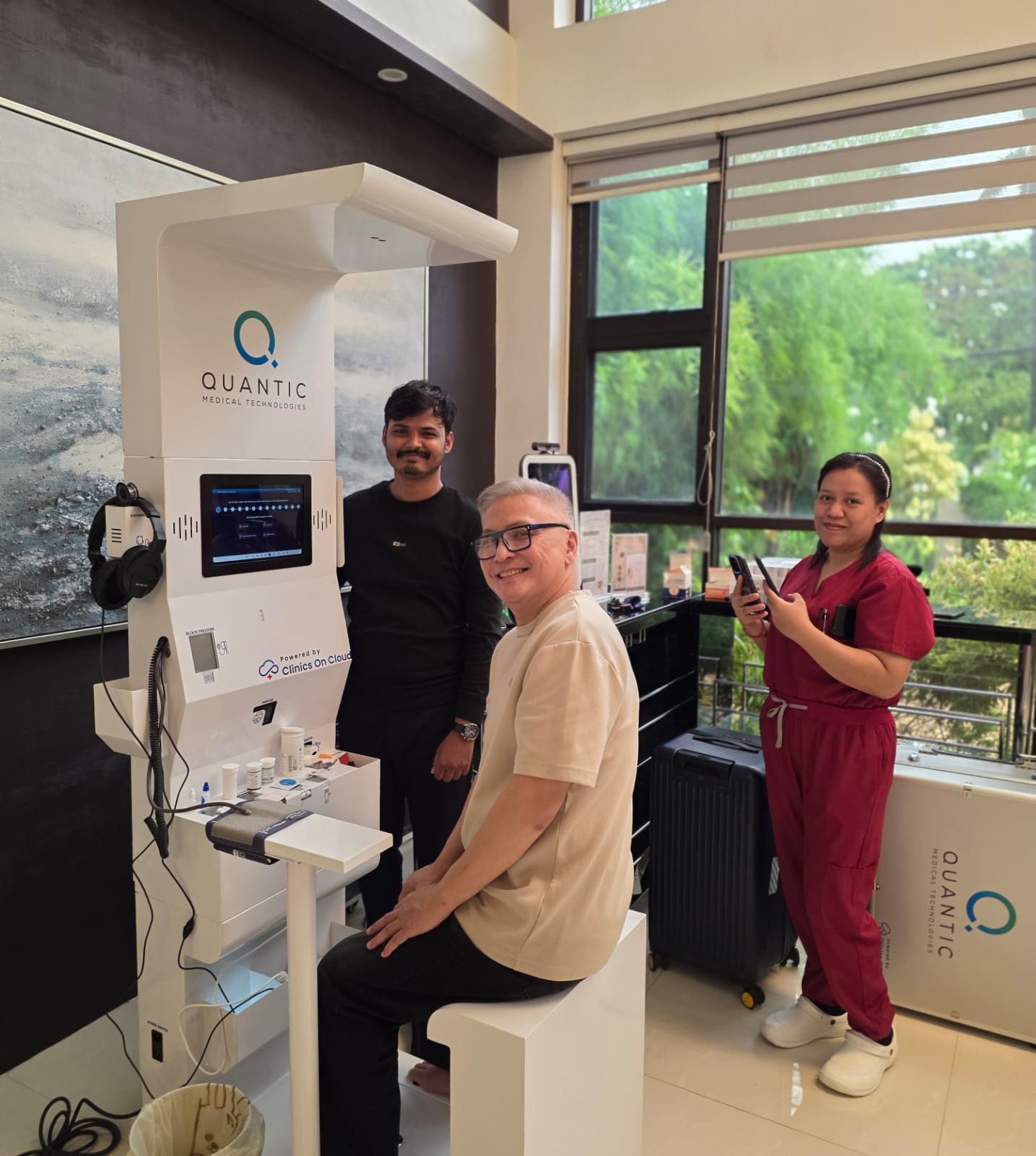 Man seated at Quantic health kiosk with blood pressure cuff, two colleagues standing beside it near a window.